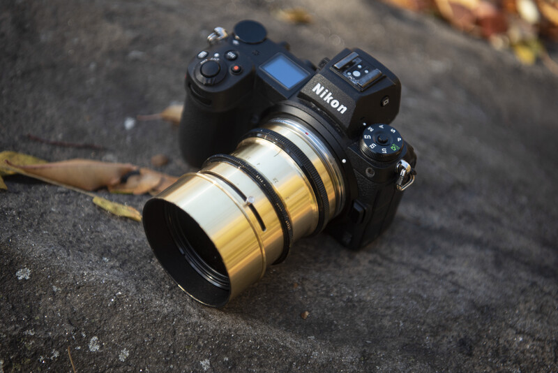 Nikon mirrorless camera with brass-colored telephoto lens on textured pavement with autumn leaves.