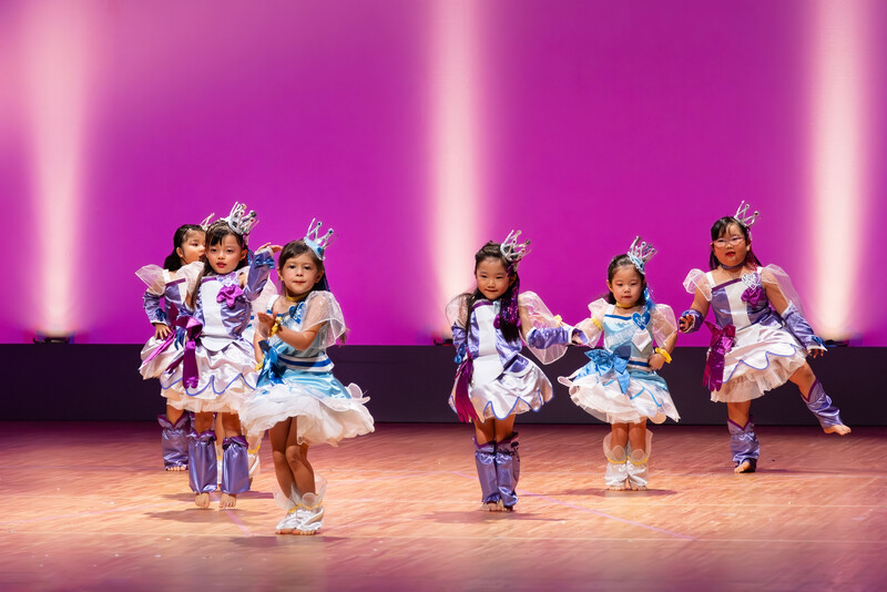 Young performers in colorful costumes dance on stage under bright pink and purple backlighting.