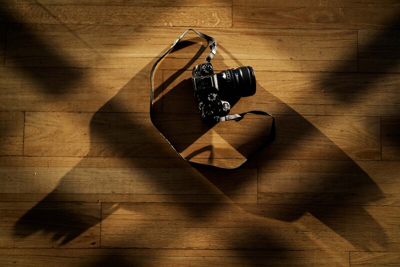 Professional camera with brown leather strap on wooden floor with dramatic window light creating geometric shadows.