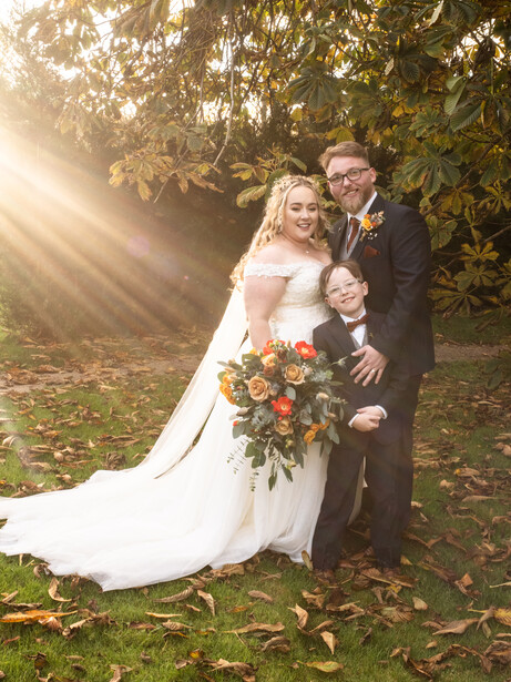 Bride and groom posing together outdoors under tree canopy with dramatic golden sunlight filtering through leaves.