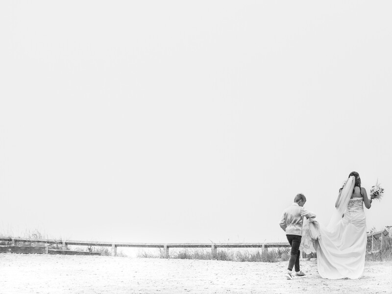Bride in flowing white dress walking with groom across a barren field near a rustic fence.