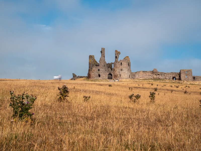 Ruined stone castle with multiple towers standing in an open, arid grassland under partly cloudy sky.