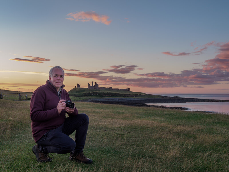 Photographer crouching in grassy field at sunset with distant castle silhouette across water.