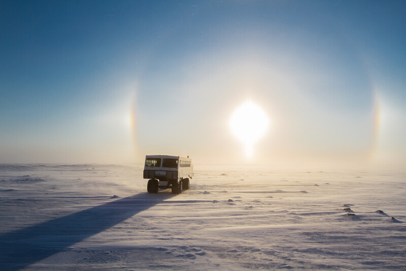Truck driving across a frozen landscape with a bright sun and sun dogs creating halos in a pale sky.
