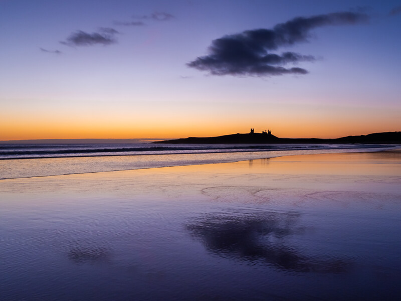 Tranquil beach at twilight with wet sand reflecting purple and golden sky above dark clouds.