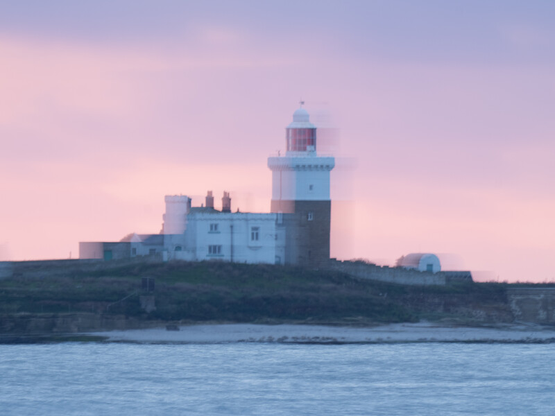 Lighthouse with attached keeper's house on rocky coastal headland at twilight with pink sky.