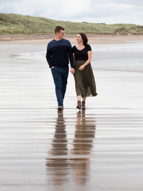 Couple walking together on a wet beach with sand dunes in the background.