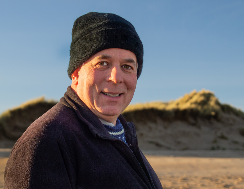 Man wearing a dark beanie and navy sweater smiling at camera on a beach with sand dunes in background.