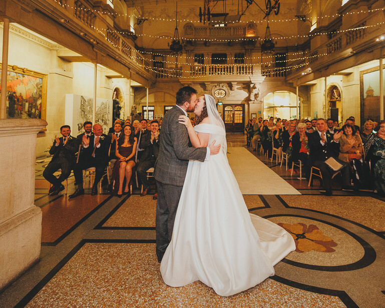 Bride and groom kiss during their first dance in an ornate, symmetrical chapel with guests seated on both sides.