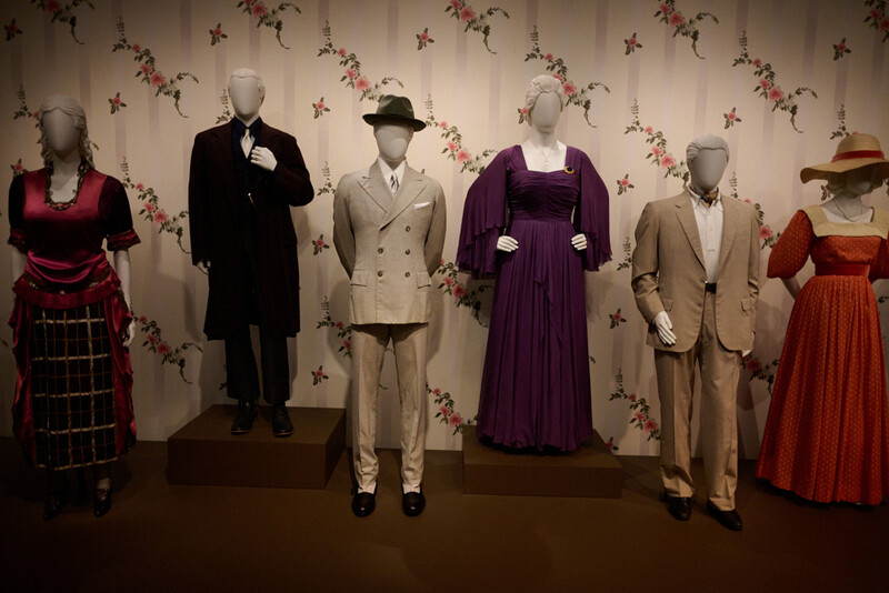 Six mannequins displaying vintage clothing from different eras against a decorative floral wallpaper backdrop.