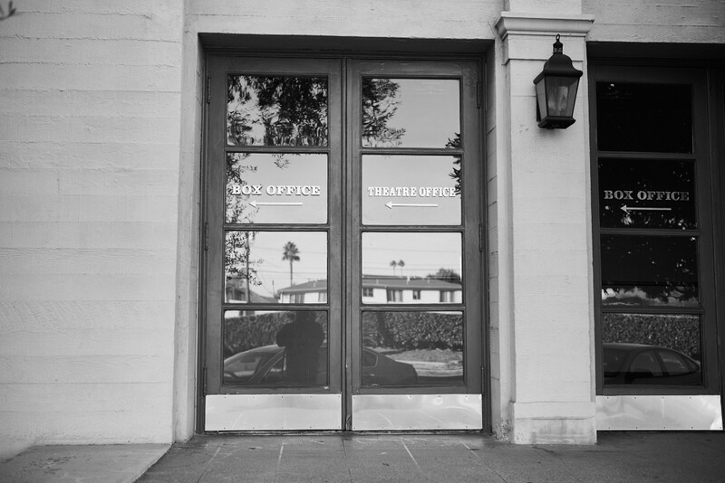 Black and white photograph of a theater entrance with glass doors and signage reflected in storefront windows.
