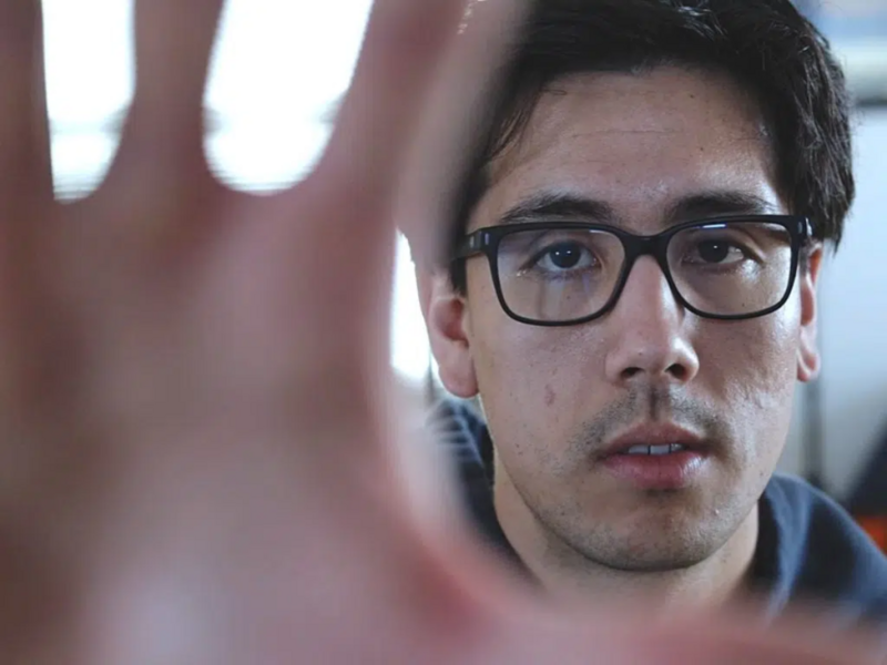 Man wearing glasses with hand raised toward camera in close-up portrait.