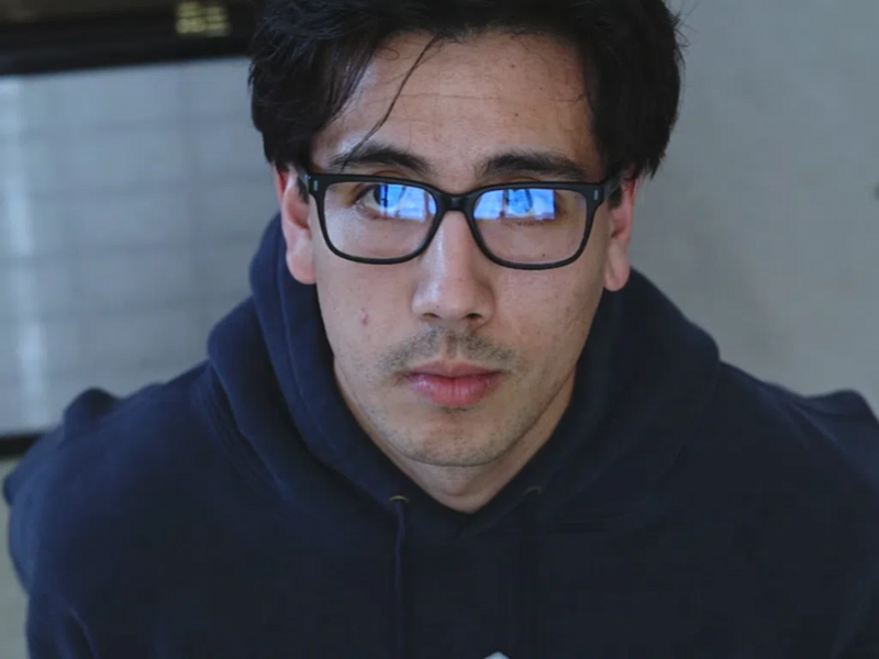 Portrait of a person wearing glasses with blue-tinted lenses and a dark hoodie against a neutral background.