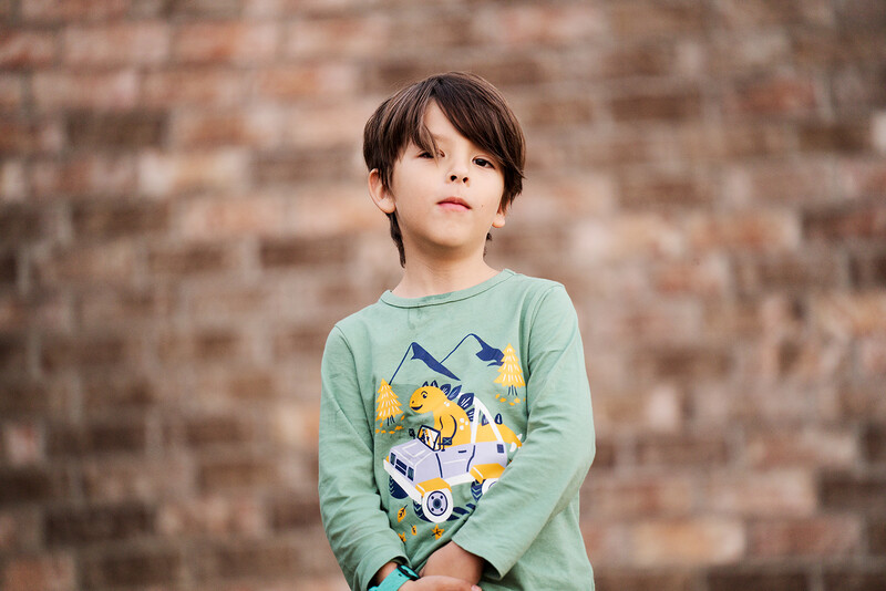 Young child in mint green long-sleeve shirt with yellow and blue graphic print against brick wall.