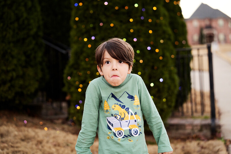 Young child in sage green long-sleeve shirt standing outdoors with blurred holiday lights and evergreen trees in background.
