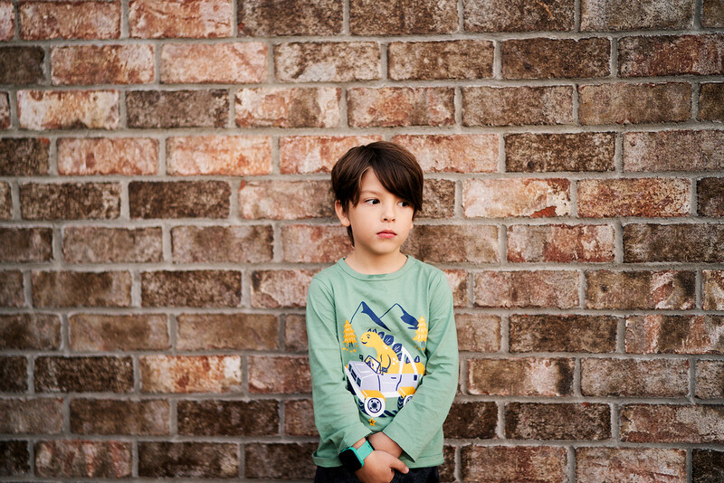 Young child in mint green graphic t-shirt standing against weathered brick wall, looking upward.