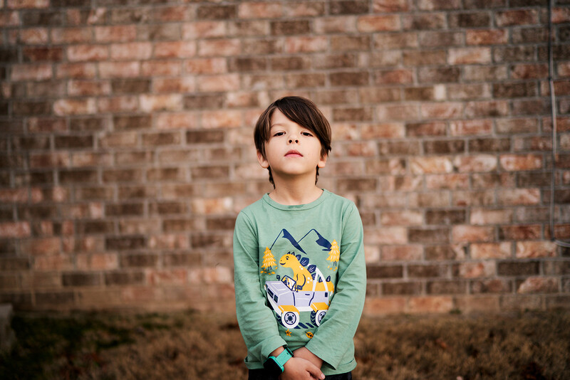 Young child in sage green long-sleeve shirt with graphic design standing against weathered brick wall, looking upward.