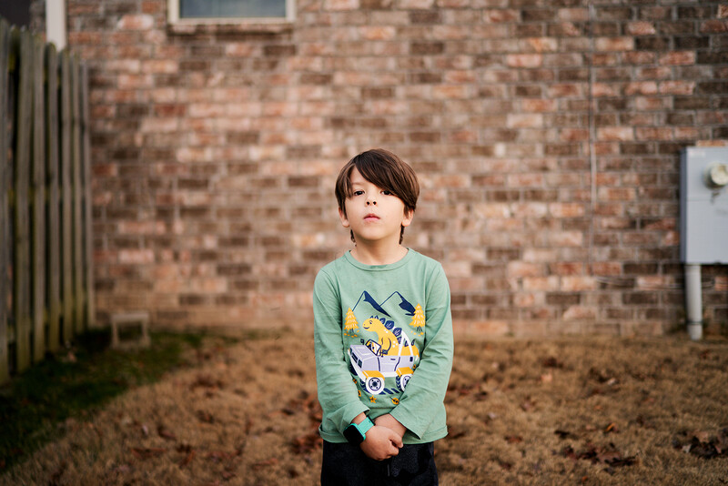 Young child in green long-sleeve shirt standing in barren yard against brick wall.