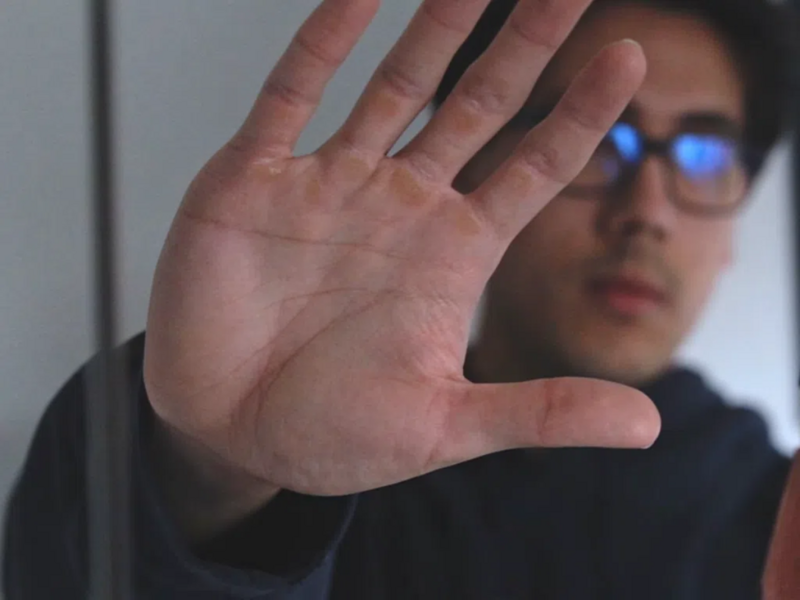Person wearing blue-tinted glasses with hand raised in a stop gesture toward the camera.