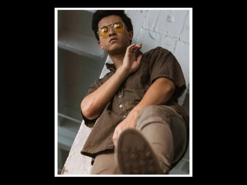Man wearing glasses and brown shirt seated indoors, hand near face in thoughtful pose.