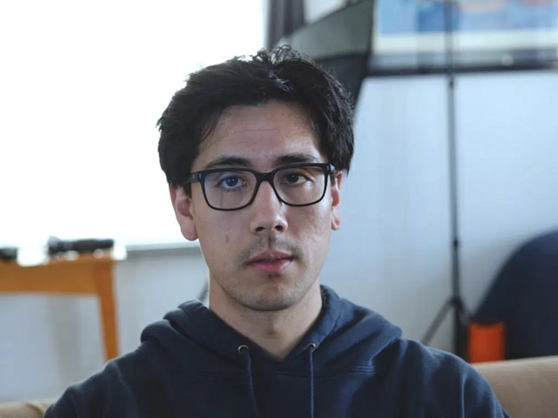 Man wearing glasses and dark hoodie in indoor setting with window background.