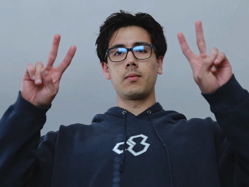 Person wearing glasses and navy hoodie making peace signs with both hands against neutral gray backdrop.
