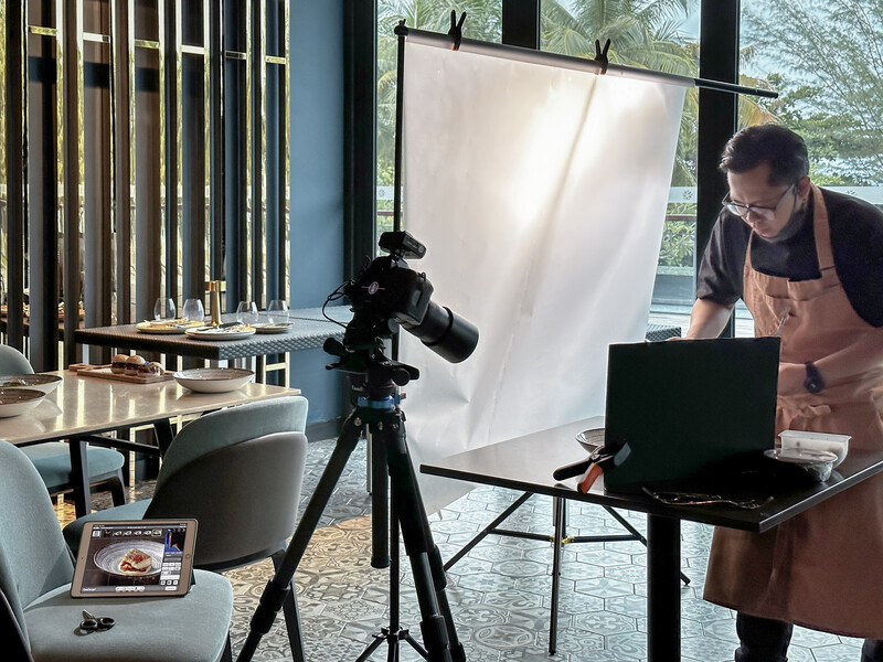 Photographer working at a laptop next to a camera on tripod with diffusion panel in bright studio space.
