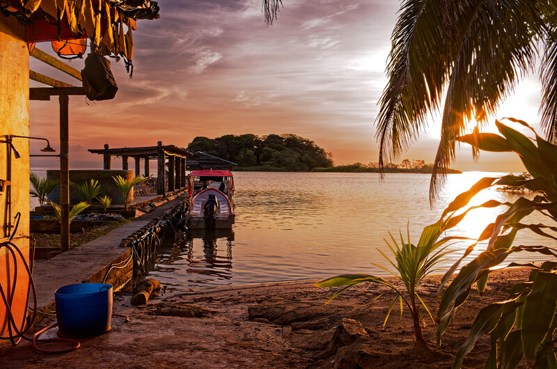 Wooden dock and beach at sunset with boats, palm trees, and calm water under golden sky.
