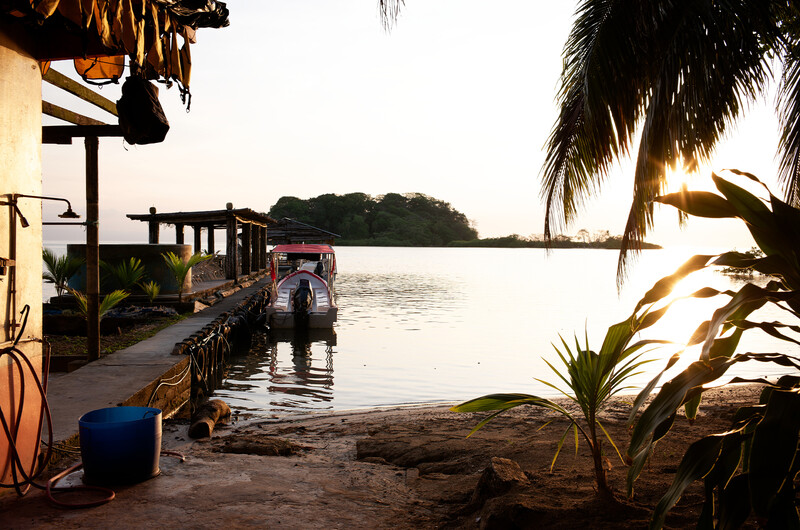 Wooden dock extending into calm water at sunset with palm trees and structures on shore.