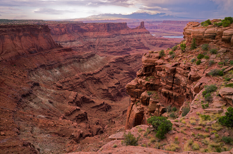Vast desert canyon landscape with layered red rock formations and distant mountains under cloudy sky.