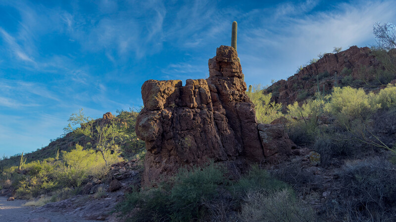 Distinctive rock formation with tall spire rising from desert hillside dotted with sparse vegetation.