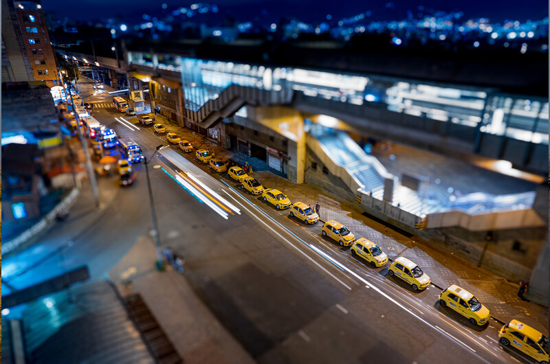 Row of yellow taxis lined up on a street at night with tilt-shift effect creating miniature appearance.