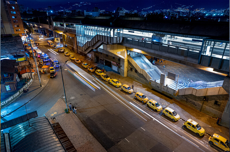 Row of yellow taxis lined up along an urban street at night beneath elevated highway infrastructure.