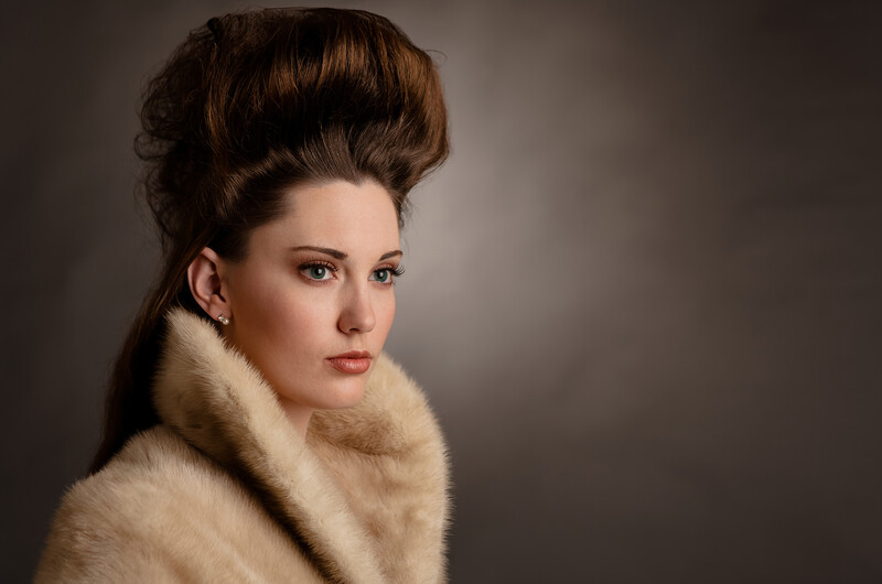 Woman with styled updo hairstyle wearing a cream-colored fur stole, photographed against a neutral gray background.