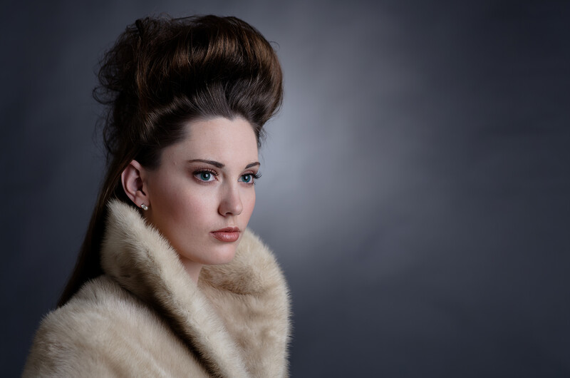 Woman with styled dark hair and pale makeup wearing a cream-colored fur collar against a neutral gray background.