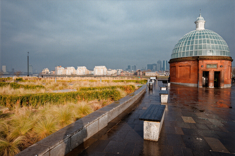 Red brick building with domed glass roof overlooking wet plaza and overgrown wetland with city skyline beyond.