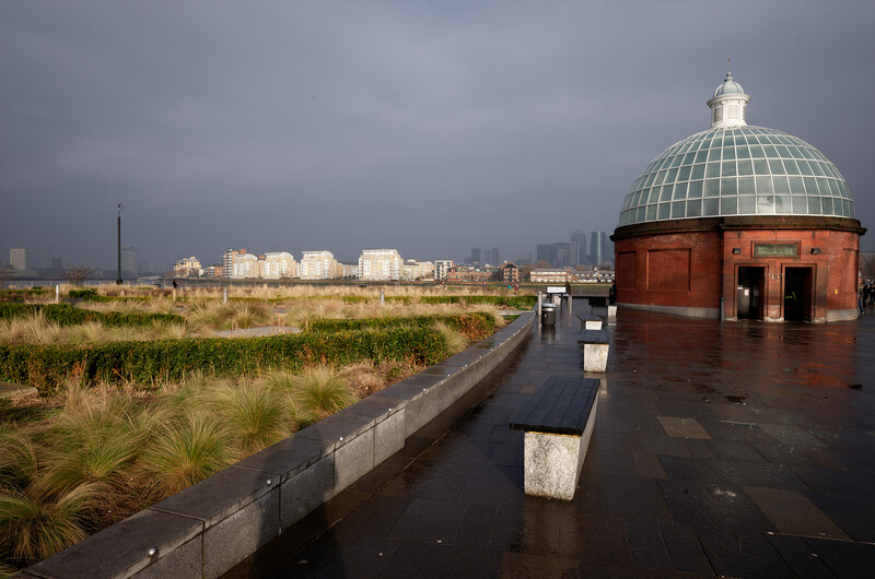 Red brick rotunda with domed glass roof beside wet plaza and grassy wetland under overcast sky.