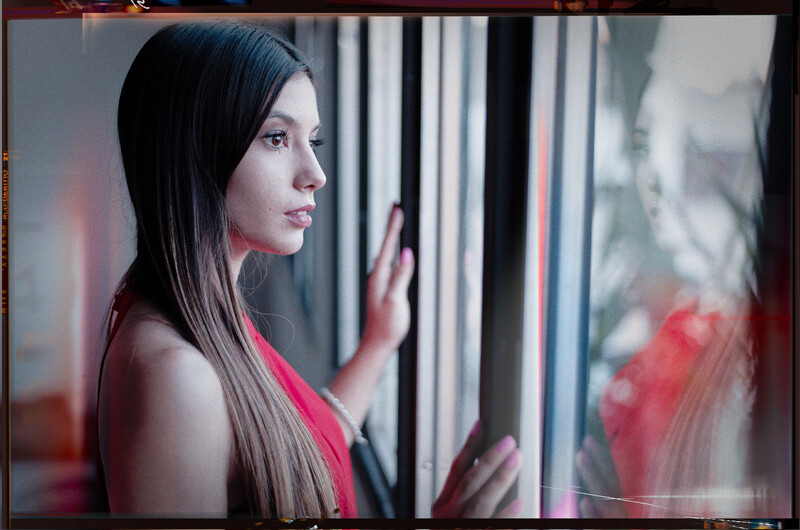 Woman with long dark hair touching a window, wearing pink, shot in profile with dramatic lighting.