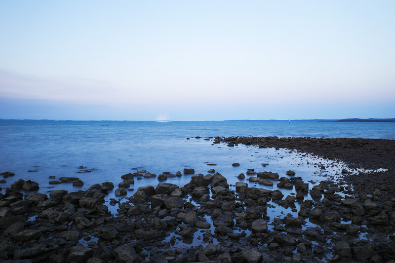 Rocky shoreline with tide pools at dusk, calm blue water and distant coastline under twilight sky.