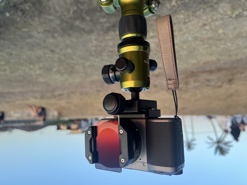 Panoramic camera mounted on a ball head tripod positioned on a beach with palm trees visible in the background.