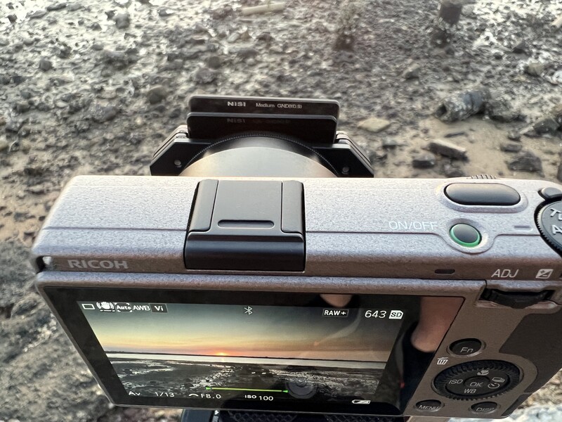 Ricoh compact camera displaying a landscape photo on its rear LCD screen, positioned on rocky ground.