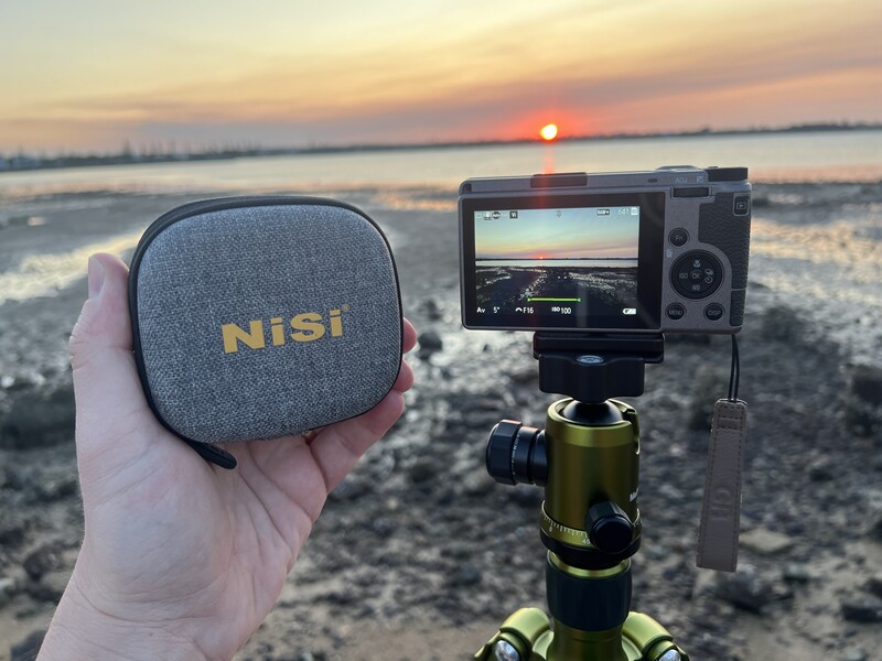 Hand holding NiSi filter case with camera on tripod during golden hour sunset over water.