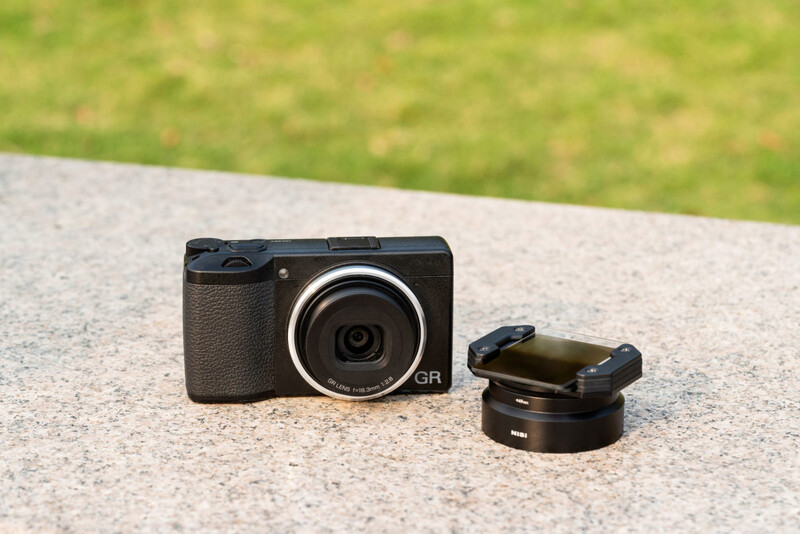 Black compact camera with textured grip and detached lens converter on concrete surface.