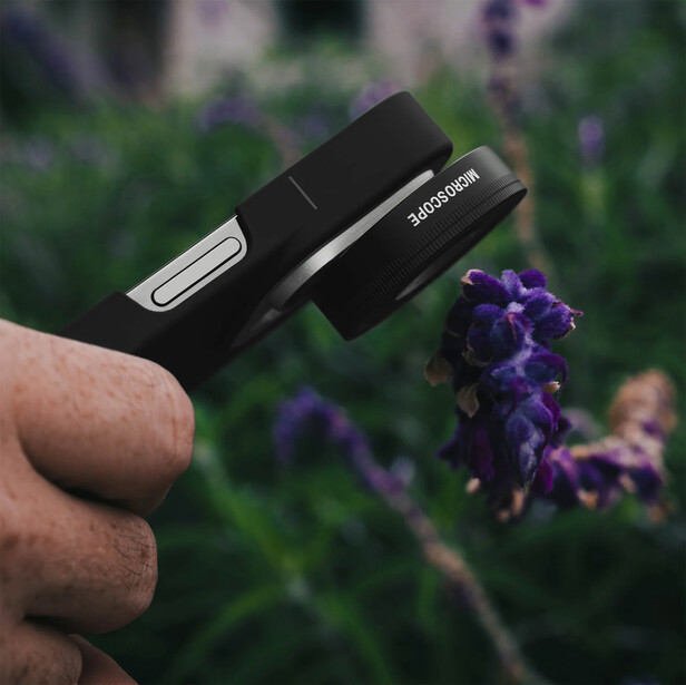 Hand holding two black camera lens caps against a blurred purple flower background.