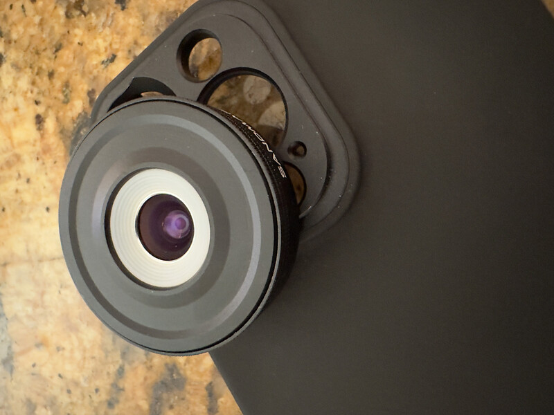 Wide-angle lens attachment mounted on smartphone camera with gray casing and purple glass element.