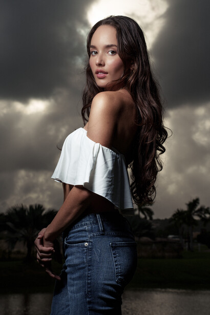 Woman with long dark hair wearing white off-shoulder top and jeans, posed against dramatic cloudy sky.