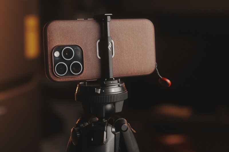Rose-gold iPhone with triple camera setup mounted on a tripod ball head against dark background.