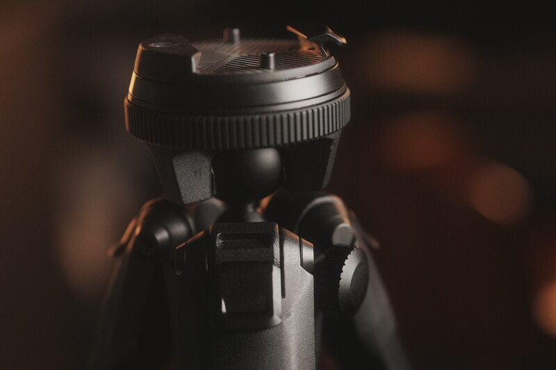 Close-up of a camera tripod head with ball joint and adjustable handles against a blurred warm-toned background.