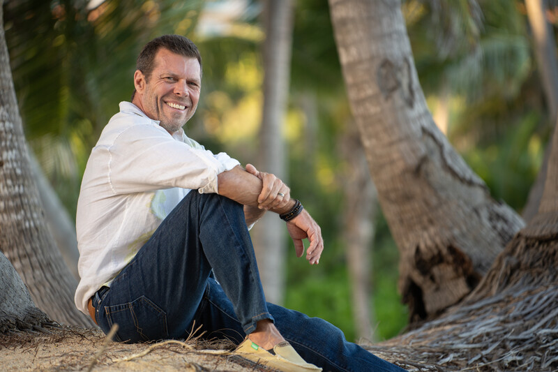 Man in white shirt and jeans sitting on sandy ground between palm tree trunks, smiling at camera.