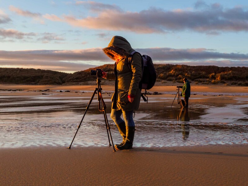 Photographer with tripod and camera on wet beach sand at golden hour, with another person visible in the background.